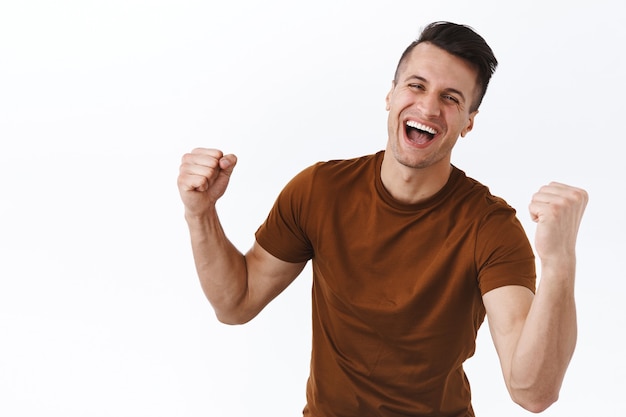 Ritratto di un ragazzo atletico felice e trionfante con bicipiti, mani forti, pompa a pugno e gridando sì, sorridente celebrando la vittoria, raggiungere l'obiettivo o il successo, diventare campione, stare in piedi sul muro bianco