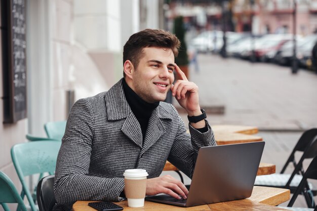 Ritratto di un ragazzo di successo che lavora con un laptop argentato in un caffè di strada, pensando al business o chiacchierando con un amico