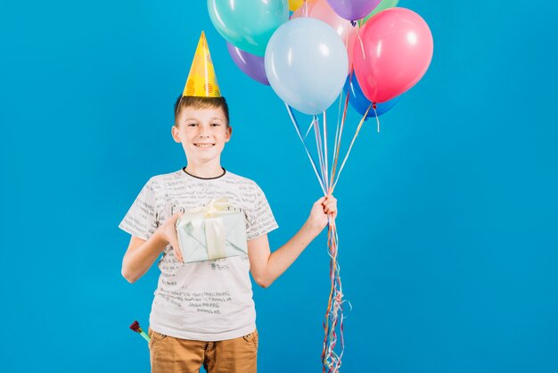 Ritratto di un ragazzo felice che tiene palloncini colorati e regalo di compleanno su sfondo blu