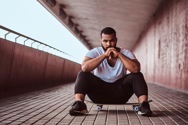 Ritratto di un ragazzo pensoso dalla pelle scura vestito con una camicia bianca e pantaloncini sportivi seduto su uno skateboard su un marciapiede sotto un ponte.