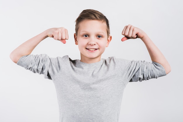 Ritratto di un ragazzo sorridente che serra il suo pugno che guarda alla macchina fotografica che si leva in piedi contro la priorità bassa grigia