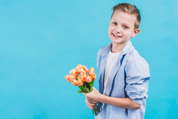 Ritratto di un ragazzo sorridente che tiene bei tulipani freschi in mano in piedi contro la parete blu