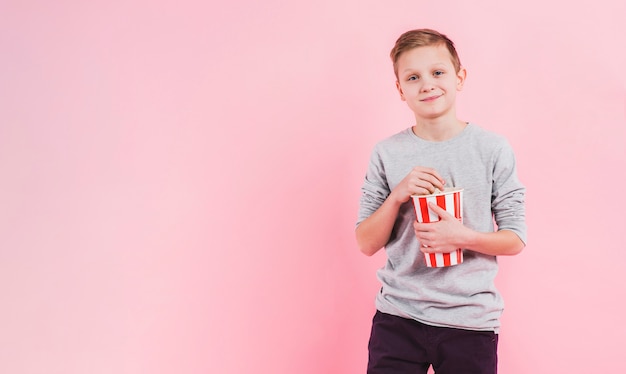 Ritratto di un secchio sorridente del popcorn della tenuta del ragazzo contro fondo rosa