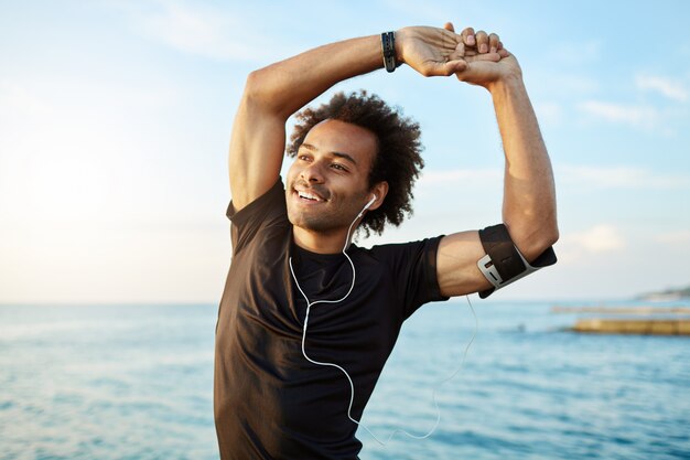 Ritratto di un sorridente afro-americano sportivo che allunga le braccia muscolose prima dell'allenamento in riva al mare, utilizzando l'app musicale sul suo smartphone.