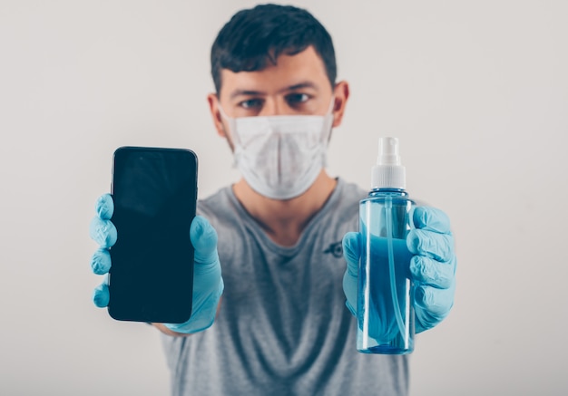 Ritratto di un uomo a sfondo chiaro in possesso di un telefono e disinfettante per le mani in guanti e maschera medica