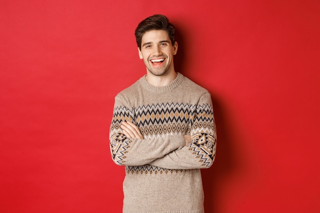 Ritratto di un uomo allegro e attraente con un maglione natalizio, che ride e sorride, celebra il nuovo anno e le vacanze invernali, in piedi su sfondo rosso