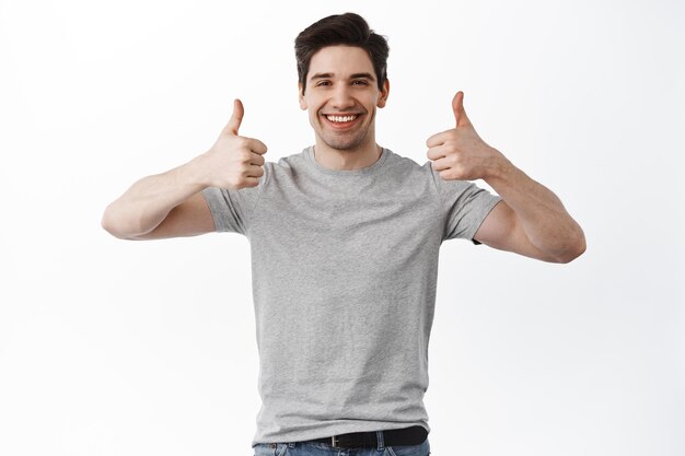 Ritratto di un uomo allegro in abbigliamento di base che sorride e mostra i pollici alla telecamera isolata su sfondo bianco