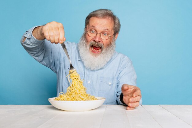 Ritratto di un uomo anziano che mangia una deliziosa cena italiana, mangiando pasta isolata su sfondo blu