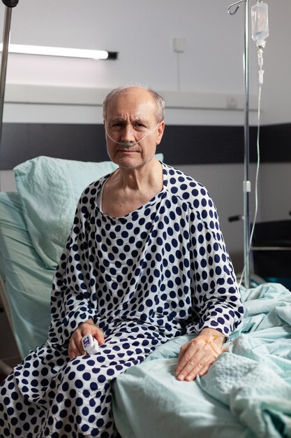 Ritratto di un uomo anziano triste e malato seduto sul bordo del letto d'ospedale con flebo iv attaccato e che respira con l'aiuto della maschera di ossigeno, guardando la telecamera.