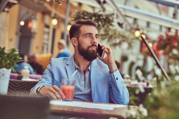 Ritratto di un uomo d'affari barbuto alla moda con un taglio di capelli alla moda, parlando al telefono, beve un bicchiere di succo, seduto in un caffè all'aperto.