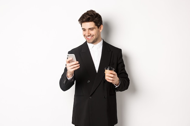 Ritratto di un uomo d'affari bello e sicuro in abito nero, che beve caffè e usa il telefono cellulare, sorride soddisfatto, in piedi su sfondo bianco