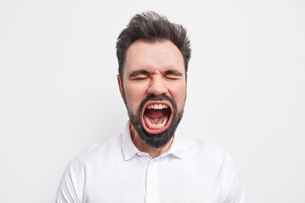 Ritratto di un uomo europeo pazzo barbuto emotivo tiene la bocca spalancata e chiude gli occhi ha una barba folta vestita di camicia