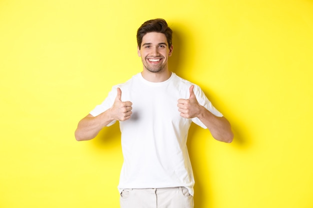 Ritratto di un uomo felice che mostra il pollice in su in segno di approvazione, come qualcosa o d'accordo, in piedi su sfondo giallo