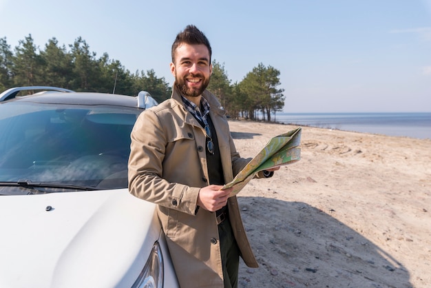 Ritratto di un viaggiatore maschio che sta vicino alla mappa della tenuta dell&#39;automobile in mano che esamina macchina fotografica