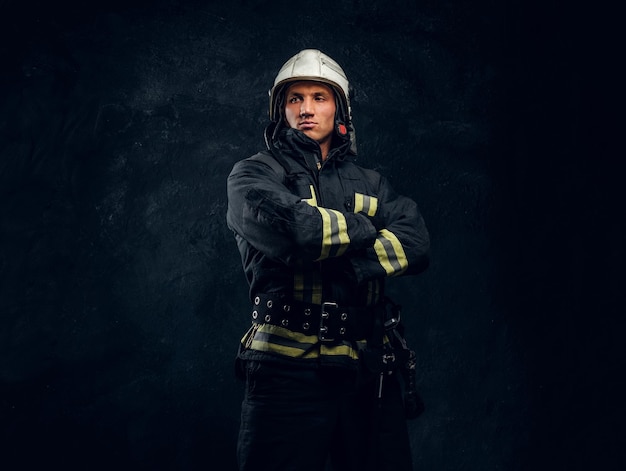 Ritratto di un vigile del fuoco in uniforme e casco sta con le mani incrociate, guardando lateralmente con uno sguardo sicuro. Foto dello studio contro una parete strutturata scura