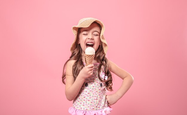 Ritratto di una bambina allegra in un cappello estivo con gelato in mano, su uno sfondo colorato di rosa, concetto di estate