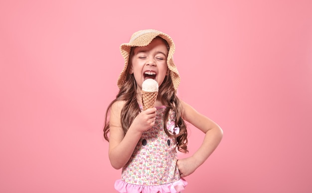 Ritratto di una bambina allegra in un cappello estivo con gelato in mano, su uno sfondo colorato di rosa, concetto di estate