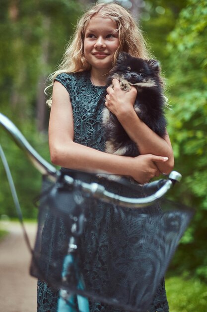 Ritratto di una bambina bionda sorridente in un abito casual, tiene un simpatico cane spitz. Andare in bicicletta in un parco.