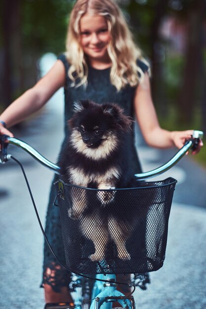 Ritratto di una bambina bionda sorridente in un abito casual, tiene un simpatico cane spitz. Andare in bicicletta in un parco.