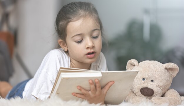 Ritratto di una bambina carina che legge un libro a casa, sdraiata sul pavimento con il suo giocattolo preferito.