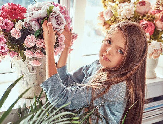 Ritratto di una bambina carina con lunghi capelli castani e sguardo penetrante che indossa un abito elegante, in posa con fiori contro la finestra di casa.
