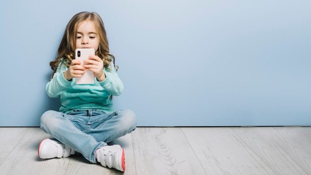 Ritratto di una bambina seduta sul pavimento in legno guardando smartphone