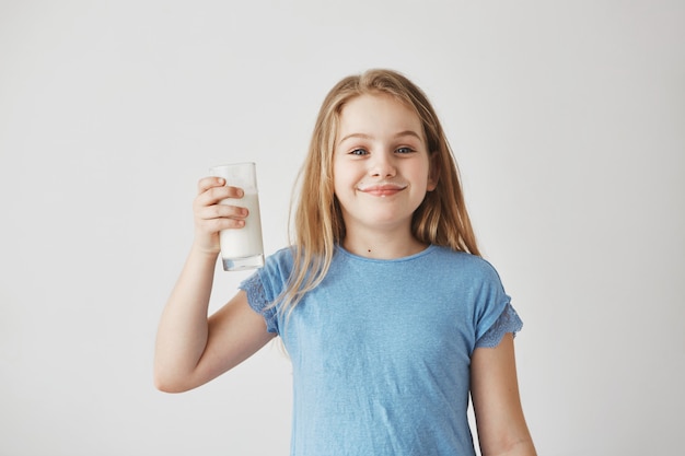 Ritratto di una bella bambina con i capelli lunghi biondi e gli occhi azzurri, sorridente con in mano un bicchiere di latte, essendo felice dopo aver bevuto preferito.