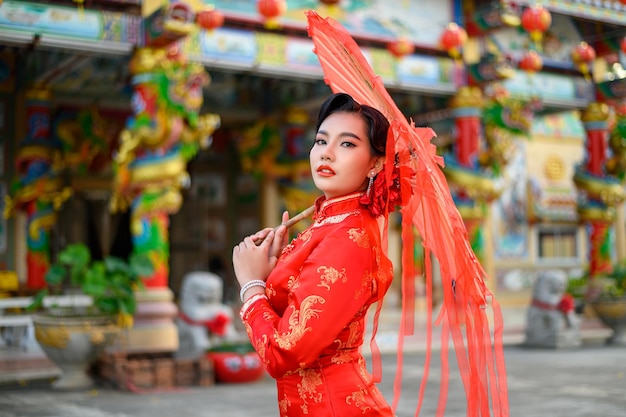 Ritratto di una bella donna asiatica che indossa un cheongsam sorridente e posa con un ombrello rosso di carta al santuario il capodanno cinese