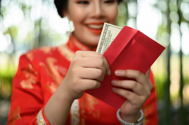 Ritratto di una bella donna asiatica in un cheongsam cinese che tiene in mano buste e ventaglio di banconote in dollari con happinese nella foresta di bambù