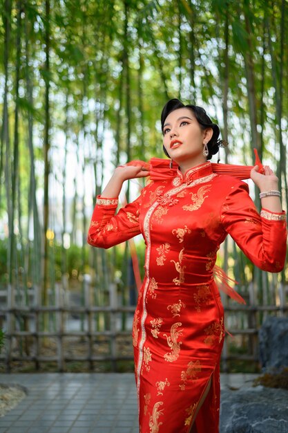 Ritratto di una bella donna asiatica in un cheongsam cinese in posa con un bellissimo ombrello di carta rosso sulla foresta di bambù, copia spazio