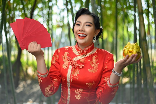 Ritratto di una bella donna asiatica in un cheongsam cinese in posa con un ventaglio di buste rosse e un salvadanaio dorato nella foresta di bambù