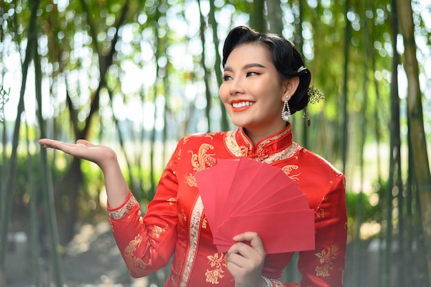 Ritratto di una bella donna asiatica in un cheongsam cinese tiene un ventaglio di buste rosse e apre le palme nella foresta di bambù