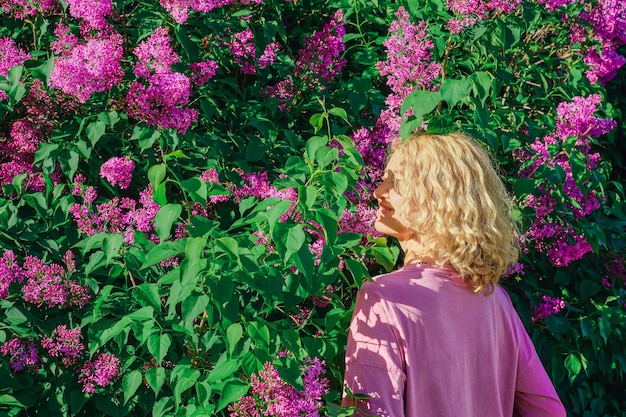 Ritratto di una bella donna bionda adulta con lunghi capelli ondulati si trova in un giardino fiorito in cespugli di lillà Primavera una donna adulta gode di primavera la natura riposa in un parco di lillà di campagna