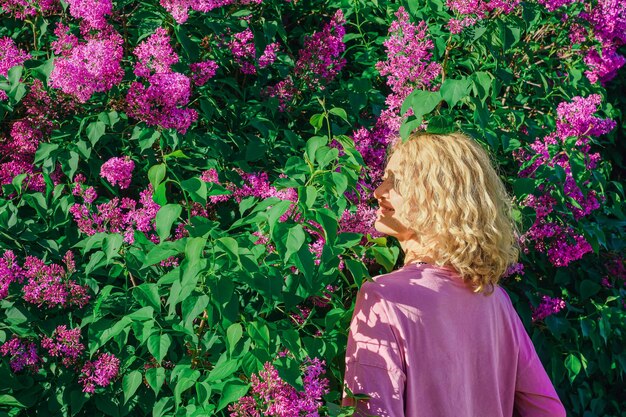 Ritratto di una bella donna bionda adulta con lunghi capelli ondulati si trova in un giardino fiorito in cespugli di lillà Primavera una donna adulta gode di primavera la natura riposa in un parco di lillà di campagna