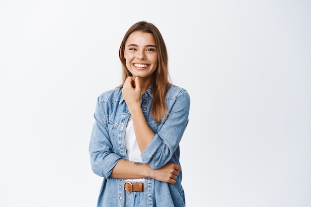 Ritratto di una bella donna sorridente che si tocca il viso con un trucco naturale e sembra allegra davanti, in piedi contro il muro bianco