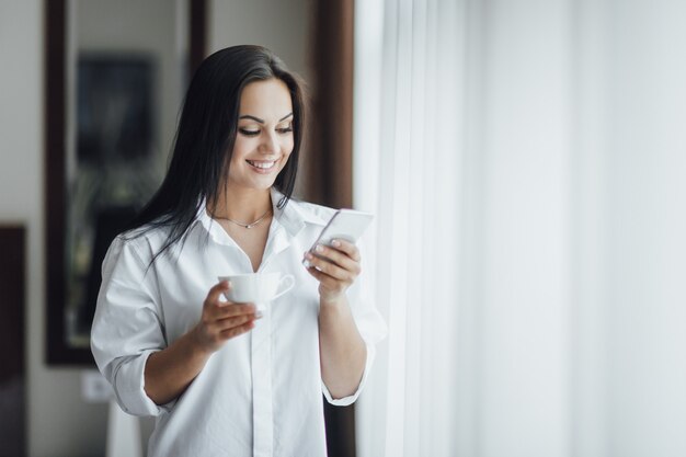 Ritratto di una bella ragazza bruna felice al mattino con caffè e un telefono vicino alla finestra.