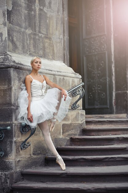 Ritratto di una bellissima giovane ballerina bionda in abito bianco in piedi con grazia sulle scale di un vecchio castello.