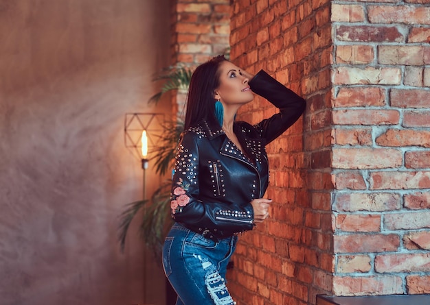 Ritratto di una bruna sensuale sexy in posa in giacca di pelle alla moda e jeans appoggiati a un muro di mattoni.