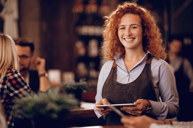 Ritratto di una cameriera rossa felice che tiene il touchpad mentre è in piedi in un pub e guarda la fotocamera