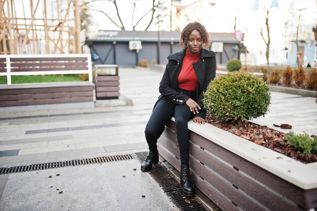 Ritratto di una donna africana dai capelli ricci che indossa un cappotto nero alla moda e un dolcevita rosso in posa all'aperto