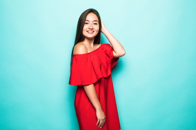 Ritratto di una donna asiatica sorridente in vestito rosso che sta esaminante macchina fotografica isolata sopra il fondo del turchese