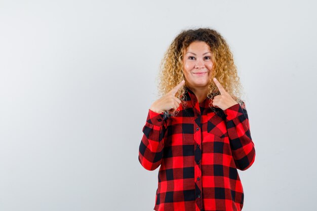 Ritratto di una donna bionda carina che indica il suo sorriso in camicia a quadri e sembra una vista frontale allegra