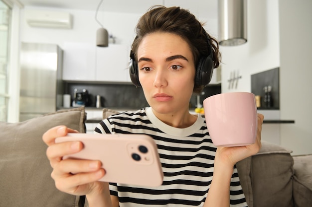 Ritratto di una donna con un viso spaventato e preoccupato che guarda un video su uno smartphone che beve tè indossando