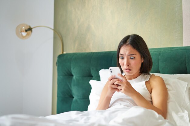 Ritratto di una donna coreana scioccata che guarda il suo telefono cellulare sdraiato a letto a leggere cattive notizie scioccanti