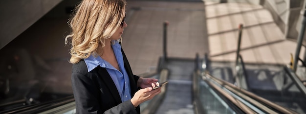 Ritratto di una donna d'affari in giacca e cravatta in piedi sulla scala mobile e che utilizza il telefono cellulare