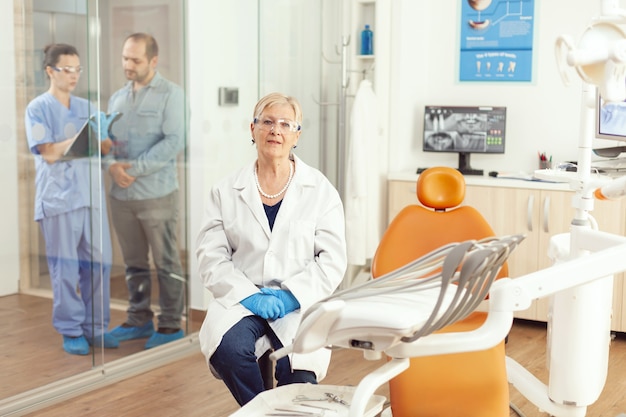 Ritratto di una donna dentista anziana sorridente in studio dentistico mentre un'infermiera medica parla con il paziente in background