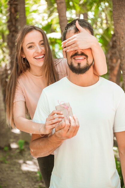 Ritratto di una donna felice che copre gli occhi del suo ragazzo e dandogli un regalo a sorpresa