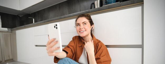 Ritratto di una donna felice e sorridente in chat video seduta sul pavimento della cucina a casa a parlare con un amico