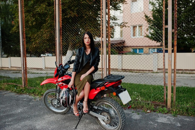 Ritratto di una donna fresca e fantastica in abito e giacca di pelle nera seduta su una bella moto rossa