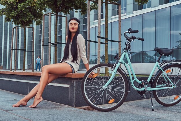 Ritratto di una donna hippie sorridente sexy che indossa una camicetta e pantaloncini in una fascia, si siede a piedi nudi su una panchina vicino a una bici da città contro un grattacielo.